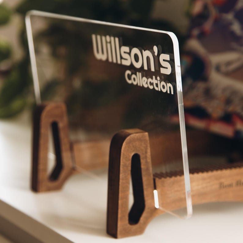 May include: A clear acrylic display stand featuring the text Willson's Collection in white lettering. The transparent panel is supported by two dark brown wooden bases with a cutout design, providing a stable structure for holding items.