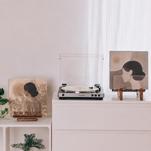 May include: A silver and black turntable sits between two wooden display stands. The left stand holds a square record sleeve with the text NOW PLAYING and an illustration of a person. The right stand holds a square record sleeve with a profile illustration.