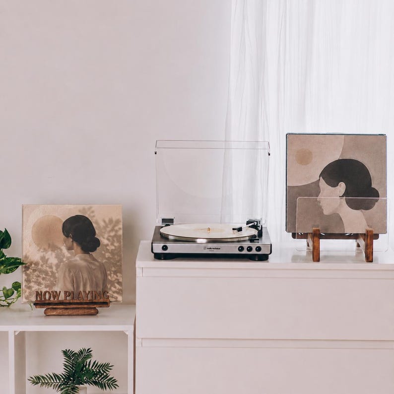 May include: A silver and black turntable sits between two wooden display stands. The left stand holds a square record sleeve with the text NOW PLAYING and an illustration of a person. The right stand holds a square record sleeve with a profile illustration.