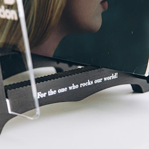May include: A black wooden display stand featuring a serrated top edge and the white printed text "For the one who rocks our world!" on its side. The stand holds a dark rectangular object with a partial image of a person's face visible above.