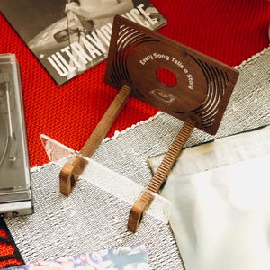 May include: A dark brown wooden display stand featuring a laser-cut design of a vinyl record and the text Every Song Tells a Story. The stand has two angled support arms and a clear acrylic base piece, designed to hold a single vinyl record sleeve.