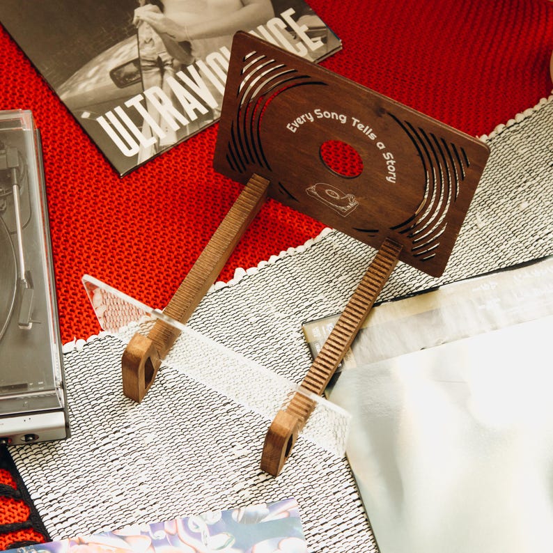 May include: A dark brown wooden display stand featuring a laser-cut design of a vinyl record and the text Every Song Tells a Story. The stand has two angled support arms and a clear acrylic base piece, designed to hold a single vinyl record sleeve.