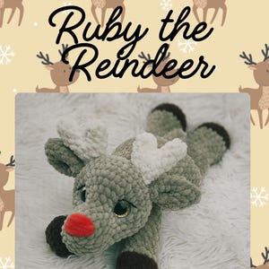 Op de afbeelding: Een handgemaakt, pluche rendier speelgoed in tinten grijs, bruin en wit, met een rode neus en grote, gouden ogen. De tekst "Ruby the Reindeer" staat boven het speelgoed. Het rendier ligt op een wit, pluizig oppervlak.