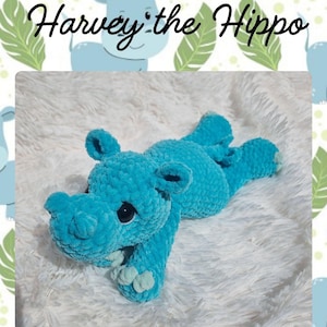 May include: A turquoise crocheted hippo toy named "Harvey the Hippo" lies on a white, fluffy surface. The hippo has black eyes and is made of soft, textured yarn. The background features a pattern of hippos and leaves.