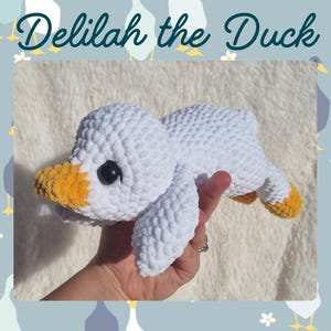 Op de afbeelding: Een handgemaakte witte gehaakte eend met een gele snavel en poten en zwarte ogen. De tekst "Delilah the Duck" staat boven de eend. De eend ligt op een zacht oppervlak.