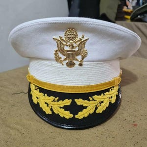 Pode incluir: Um chapéu branco de estilo militar com um emblema de águia dourada e uma trança amarela ao redor da aba. O chapéu tem uma faixa preta com folhas bordadas em ouro.