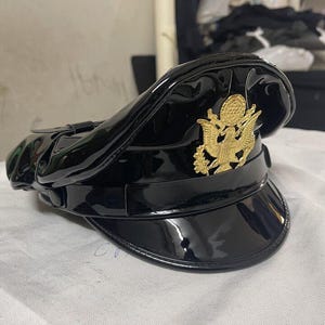 Może przedstawiać: Czarny lakierowany kapelusz w stylu wojskowym z złotym orłem. Kapelusz ma szeroki rondel i wysoki korpus.