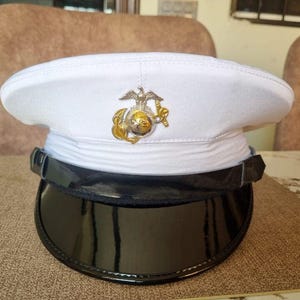 May include: White military hat with a black visor. The hat features a gold and silver emblem on the front. The hat is sitting on a brown surface.