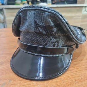 May include: A black leather cap with a snakeskin pattern. The cap has a black leather brim and a black leather band with a silver buckle.