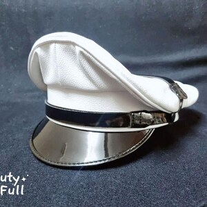 Puede incluir: Una gorra blanca de pico con una banda negra y una visera negra brillante. La gorra tiene una superficie ligeramente texturizada.