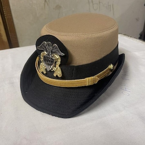 Könnte beinhalten: Eine militärische Mütze in Beige und Schwarz mit goldenem Band und Emblem. Der Hut hat eine schwarze Krempe und eine beige Krone. Ein goldenes Band ist um den Rand gewickelt, und ein silbernes und goldenes Emblem befindet sich an der Seite.