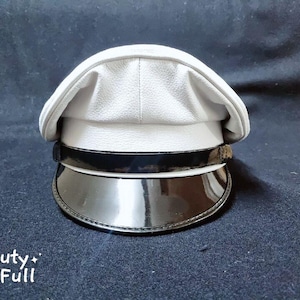 Puede incluir: Una gorra de pico de cuero blanco con una visera de charol negro. La gorra tiene una banda blanca alrededor de la visera.