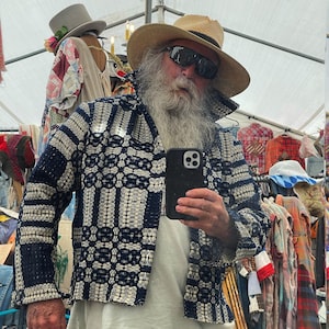 May include: A man with a long white beard and sunglasses is wearing a blue and white patterned jacket. He is wearing a straw hat and holding a black smartphone.
