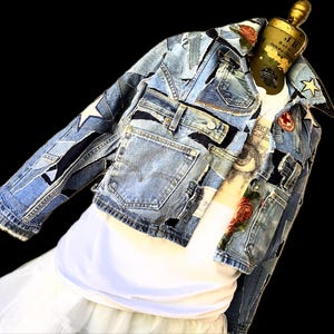 May include: A patchwork denim jacket with various shades of blue denim. The jacket features star and floral appliqués, and multiple pockets. The jacket is displayed over a white t-shirt and a white skirt. The jacket is on a mannequin.