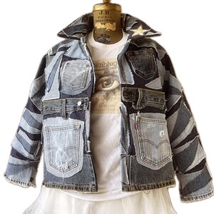 May include: A patchwork denim jacket with various shades of blue denim pieces. The jacket features multiple pockets, a collar, and a button-up front. A white t-shirt with an eye graphic is visible underneath. The jacket is displayed on a mannequin.