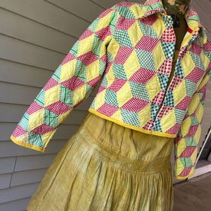 May include: A cropped quilted jacket featuring a geometric pattern of yellow, red, and green gingham diamonds. The jacket has a collar, long sleeves, and a yellow hem. Underneath is a mustard yellow skirt with a pleated design and lace trim.