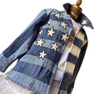 May include: A denim jacket with a patchwork design of blue denim stripes and white star appliqués. The jacket has a collared neckline and long sleeves. The jacket is displayed on a mannequin.