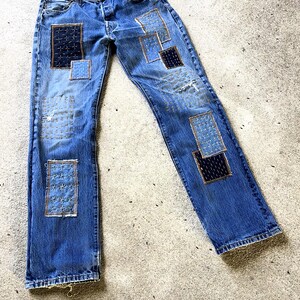 May include: Blue denim jeans with multiple patches in various shades of blue and navy. The patches are stitched on with gold thread. The jeans have a button fly and a distressed look.
