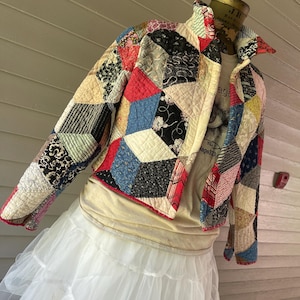 May include: A cropped jacket constructed from a patchwork of quilted fabric pieces in various colors and patterns, including black, red, blue, and cream. The jacket features a high collar, long sleeves, and a front button closure, displayed on a mannequin.
