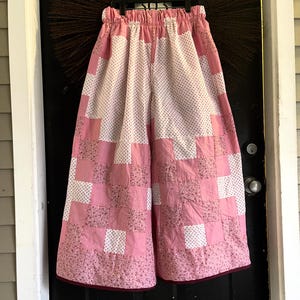 May include: Pink and white patchwork pants with an elastic waistband. The pants feature a checkerboard pattern of solid pink squares and squares with a floral print. The bottom hem is trimmed with a dark red border.