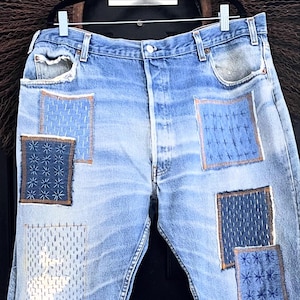 May include: Light blue denim jeans with multiple patches of various blue stitched designs. The patches are rectangular and square, with visible stitching in contrasting colors. The jeans are hanging on a hanger.