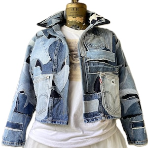 May include: A blue denim jacket made from patchwork pieces of denim. The jacket has a cropped length and a collar. The jacket is on a mannequin wearing a white t-shirt.