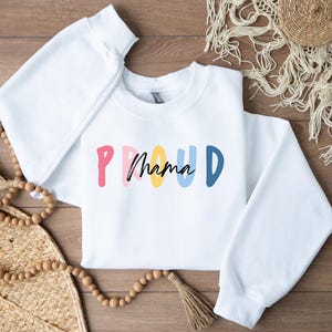 Könnte beinhalten: Weißer Pullover mit einer bunten Grafik, auf der "PROUD mama" in einem Regenbogen-Farbschema steht.
