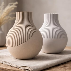 Peut inclure: Deux vases décoratifs avec un design côtelé. L'un est beige, l'autre blanc. Les deux vases ont une forme arrondie et reposent sur un tissu en lin de couleur neutre sur une table en bois. Un petit vase blanc et des plantes séchées sont en arrière-plan.