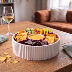May include: A round, white, fluted serving tray with multiple internal compartments. The tray contains yellow corn tortilla chips, purple blue corn tortilla chips, sliced cucumbers, and bell pepper strips, with a central bowl of yellow dip.