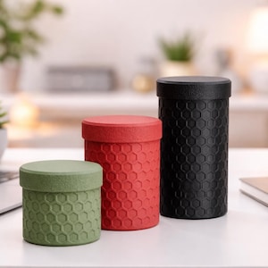 Op de afbeelding: Drie cilindrische containers in groen, rood en zwart, elk met een textuur van een honingraatpatroon. De containers zijn verschillend van grootte, waarbij de zwarte de hoogste is. Ze staan op een wit oppervlak, in de buurt van een laptop en kantoorbenodigdheden.