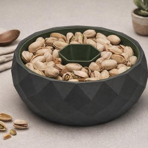 Könnte beinhalten: Eine dunkelgraue Servierschale mit geometrischem Muster, gefüllt mit Pistazien. Die Schale hat ein zentrales Fach und eine grüne Innenseite. Ein Holzlöffel und ein gefaltetes Tuch befinden sich im Hintergrund.