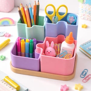 Peut inclure: Un organisateur de bureau modulaire composé de quatre compartiments en plastique aux tons pastel menthe, lavande, violet et rose, sur une base en bois clair. Il contient des crayons de couleur, des ciseaux jaunes, une figurine lapin rose, de la colle et des trombones.