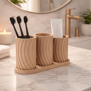 Könnte beinhalten: Ein beiges Badezimmer-Organizer-Set mit drei zylindrischen Haltern und einer rechteckigen Basis. Die Halter enthalten schwarze Zahnbürsten und eine Tube Zahnpasta. Das Set steht auf einer Marmorarbeitsplatte.