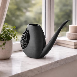 Puede incluir: Un jarrón decorativo negro en forma de pipa con un diseño calado único en el lateral. El jarrón está sobre un alféizar de mármol, con una ventana y vegetación al fondo. El jarrón tiene un pico largo y curvado.