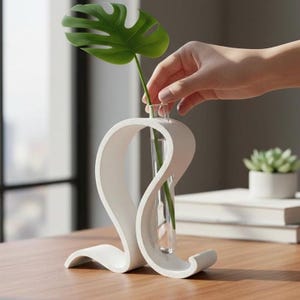 Può includere: Un vaso scultoreo bianco con un tubo di vetro trasparente per un singolo stelo. Una grande foglia verde è posta nel vaso. Il vaso ha un design unico e fluido e poggia su una superficie di legno.