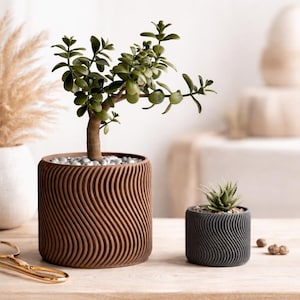 Könnte beinhalten: Zwei dekorative Pflanzgefäße mit wellenartigen Mustern. Der größere, braune Pflanztopf enthält einen kleinen Baum mit grünen Blättern und kleinen Steinen. Der kleinere, dunkelgraue Pflanztopf enthält eine Sukkulente. Beide stehen auf einem Holztisch.