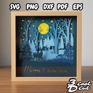 May include: A framed paper cut artwork depicting a winter forest scene. The design includes a yellow moon, stars, trees, and deer in shades of blue. The words "Merry Christmas" are cut into the paper. The frame is made of light-coloured wood.