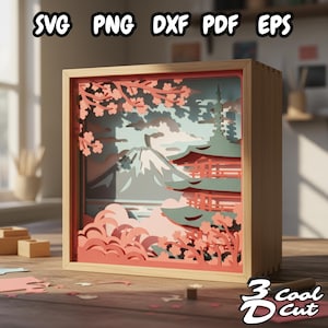 Può includere: Scatola ombra in legno con un design a strati di carta ritagliata raffigurante un paesaggio giapponese, con una montagna, una pagoda e fiori di ciliegio. Il testo "SVG PNG DXF PDF EPS" e "3 cool cut" è visibile.