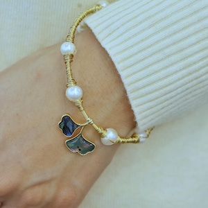 Op de afbeelding: Een armband van goudkleurig draad met witte, bolvormige parels die langs de band zijn verdeeld en twee donkere, iriserende bedels in de vorm van ginkgobladeren die in het midden hangen. Het metaaldraad is om de parels gewikkeld.