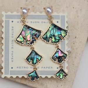 May include: A pair of dangling earrings with a gold-tone metal finish. Each earring features a pearlescent stud, a large iridescent abalone shell-shaped piece, and two smaller abalone shell-shaped pieces. The earrings are displayed on a vintage-style paper.