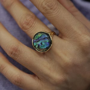 Puede incluir: Un anillo de oro con una gran piedra ovalada de nácar de abulón iridiscente. La piedra tiene un color azul, verde y morado.