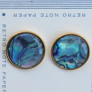 Op de afbeelding: Een paar ronde oorstekers met iriserende blauwgroene abalone schelpen, gezet in goudkleurig metaal. De oorbellen worden gepresenteerd op een witte achtergrond met de tekst "RETRO NOTE PAPER".