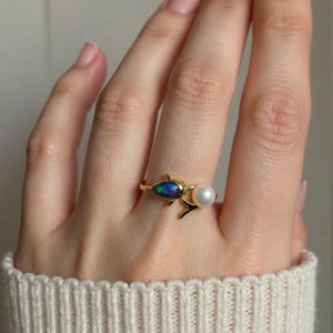 May include: A gold-toned ring featuring a fish-shaped design with a dark, iridescent opal inlay for the body and a single white spherical pearl positioned near the tail, creating a delicate and detailed piece of jewelry for the finger.