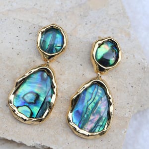 May include: A pair of drop earrings featuring two irregular, organic-shaped abalone shell inlays set in polished gold-tone metal. Each earring consists of a smaller circular top piece and a larger, elongated bottom piece with iridescent blue and green hues.