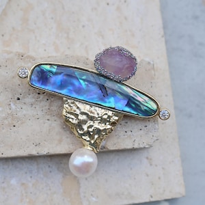 Puede incluir: Un broche dorado con una concha de abulón azul y verde, una piedra rosa y una perla. El broche tiene un diseño texturizado y abstracto.