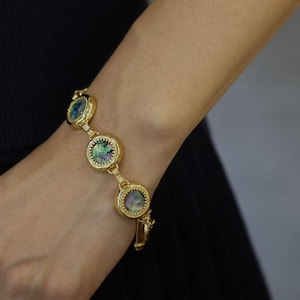 Könnte beinhalten: Ein goldfarbenes Kettenarmband mit drei kreisförmigen Gliedern, die jeweils mit einer runden, irisierenden Abalone-Muscheleinlage besetzt sind. Die Metallfassungen haben einen texturierten, perlenartigen Rand um jede Muschel und sind durch kleine Goldglieder verbunden.