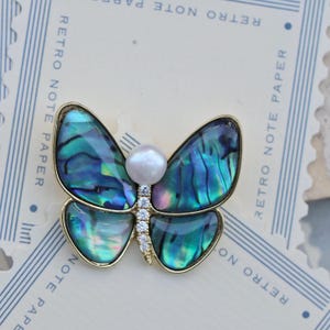 Puede incluir: Un broche de mariposa de oro con alas de concha de abulón azul y verde iridiscente y un acento de perla blanca.