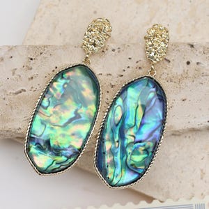 Peut inclure: Une paire de boucles d'oreilles pendantes avec des coquillages d'ormeau irisés de forme ovale, encadrés de métal doré. Les boucles d'oreilles ont des dessus texturés de couleur or. Les coquillages d'ormeau affichent des nuances de vert, de bleu et de violet.