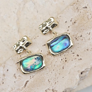 May include: A pair of gold-toned earrings. Each earring features a rectangular, iridescent abalone shell pendant with a gold frame, suspended from a textured, abstract gold stud. The abalone shell displays shades of blue, green, and purple.