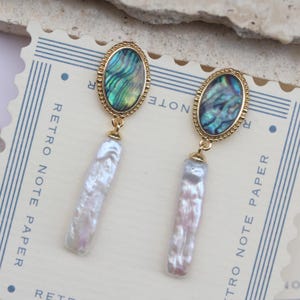 Può includere: Orecchini dorati con un pendente a goccia di perla barocca e un pendente ovale in madreperla di abalone verde e blu. Gli orecchini sono su una carta bianca con il testo "Retro Note Paper" stampato su di essa.